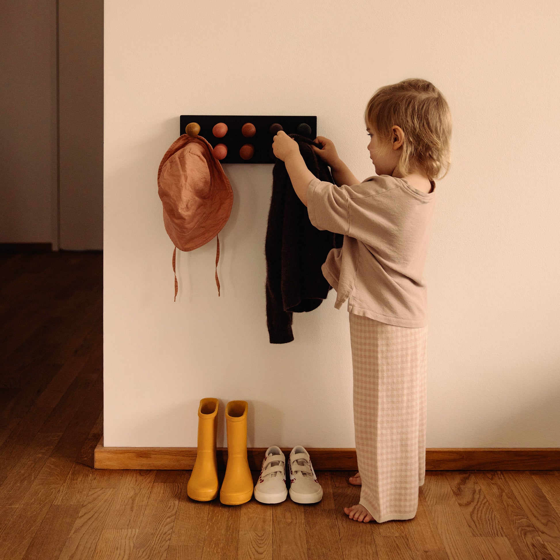 Wooden children’s wall coat rack and sorting toy in one by Woodsie Wonder – Play & Hang in Black Oak with Earth Palette, shown as a child hangs up a jumper.