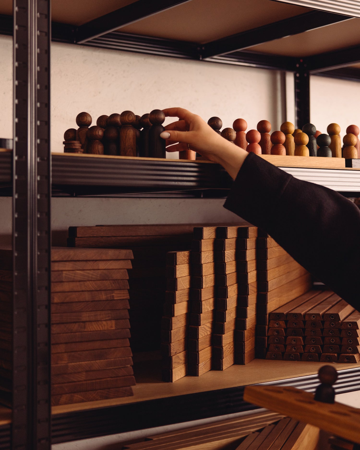 Hand selecting wooden pieces from workshop shelves beside stacked oak components used in Woodsie Wonder studio in Berlin.
